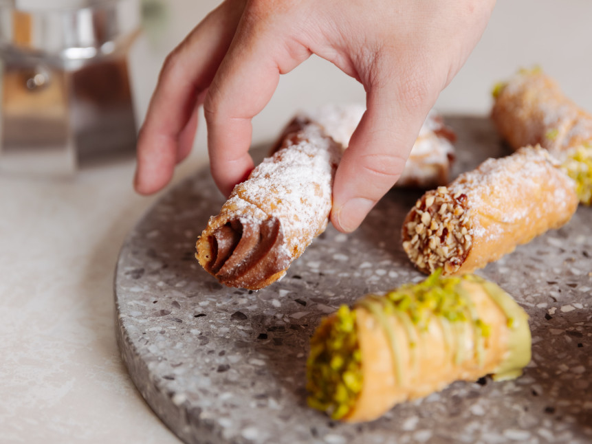 A close-up shot of a hand reaching for a chocolate-filled cannoli dusted with powdered sugar, resting on a round, grey marble tray. Several other cannoli, some topped with pistachios and others with nuts, are also on the tray.