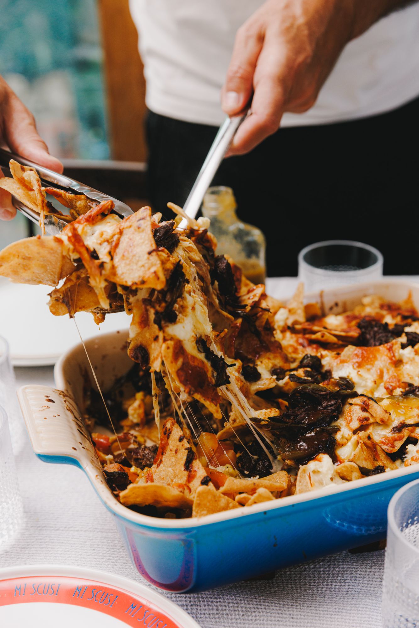 Close-up of beef cheek barbacoa nachos being served from a blue baking dish, with melted cheese stretching as tongs lift a portion, highlighting the gooey texture