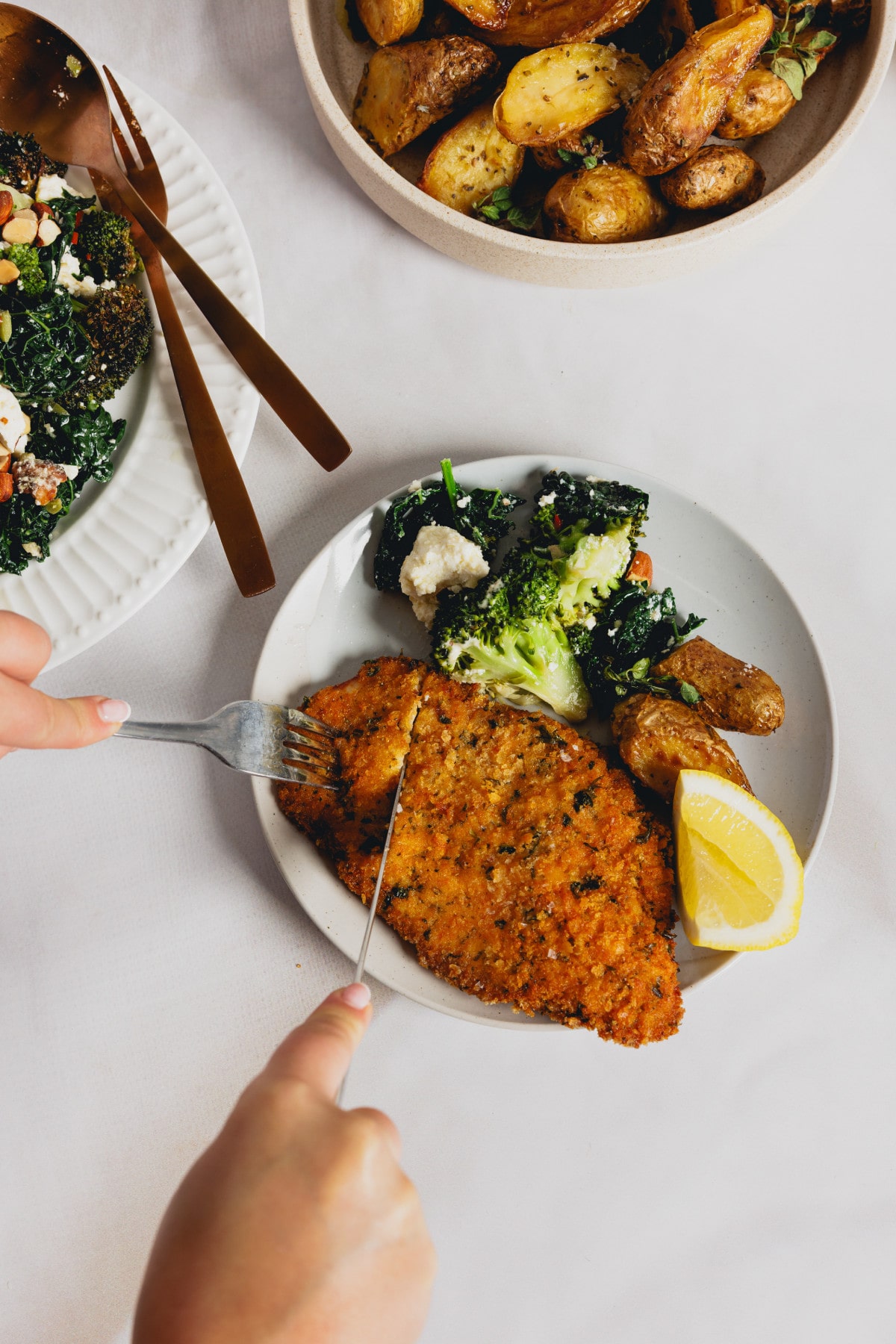 A top-down view of a person cutting into a golden-brown chicken schnitzel on a plate, served with roasted potatoes, a green salad, and a lemon wedge.