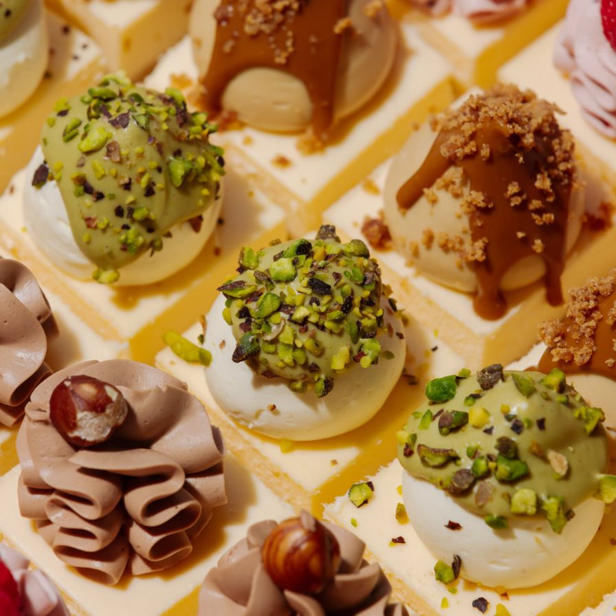 A close-up overhead view of a selection box of assorted mini patisserie cakes, including pistachio, caramel, chocolate-hazelnut, and raspberry desserts.